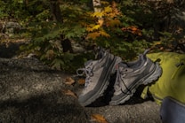 A pair of rugged hiking shoes placed on a large rock surrounded by a lush green forest. The scene includes fallen autumn leaves scattered on the rock and a portion of a green backpack visible on the right side.