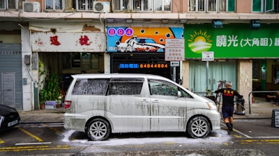 A white minivan is being washed with soap suds on a city street, presumably in front of a car wash business. A man wearing black shorts and a black shirt with the number 894 printed on the back, is seen cleaning the van. The background features three storefronts with signage in both English and Chinese, one of which has a sun logo and the other advertising the car wash service. The street and buildings appear somewhat aged with noticeable wear and tear.