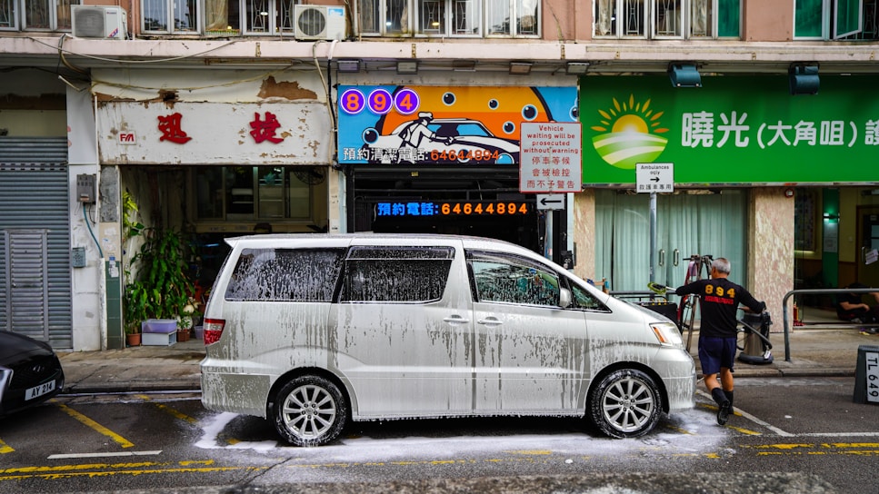 A white minivan is being washed with soap suds on a city street, presumably in front of a car wash business. A man wearing black shorts and a black shirt with the number 894 printed on the back, is seen cleaning the van. The background features three storefronts with signage in both English and Chinese, one of which has a sun logo and the other advertising the car wash service. The street and buildings appear somewhat aged with noticeable wear and tear.