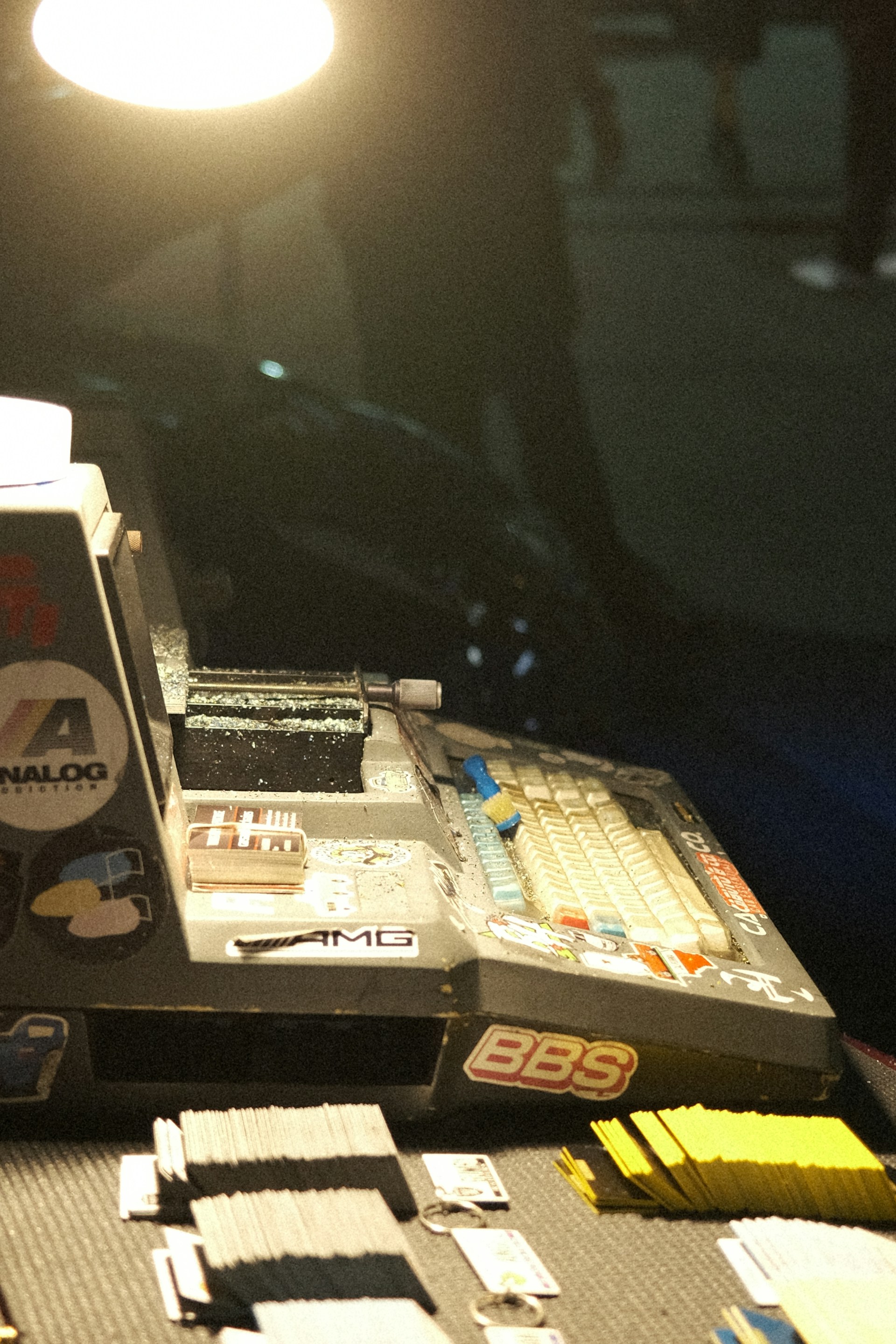 An illuminated scene featuring a retro computer setup with a keyboard, various stickers, and cards spread on a table under a bright light. The light highlights dust particles and creates a nostalgic atmosphere. The background is dim, with shadowy figures barely visible.