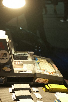 An illuminated scene featuring a retro computer setup with a keyboard, various stickers, and cards spread on a table under a bright light. The light highlights dust particles and creates a nostalgic atmosphere. The background is dim, with shadowy figures barely visible.