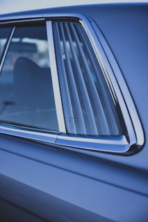 A close-up view of the side window of a car, featuring sleek chrome trim and a faint reflection visible on the glass. The car door and window have a glossy blue finish, and the background is slightly out of focus, highlighting the vehicle's features.
