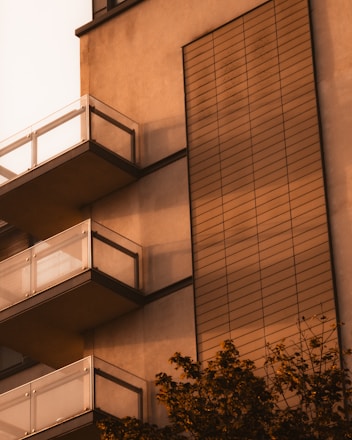 A modern building facade with several glass-panel balconies and horizontal siding. The exterior is bathed in warm, golden light, possibly from a setting sun. Foliage from a nearby tree is visible at the bottom right corner.
