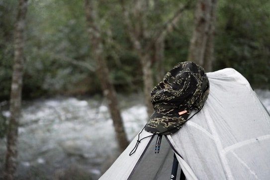 A camouflaged hat is placed atop a light gray tent in a wooded area with a river visible in the background. The scene conveys a sense of outdoor adventure and camping.