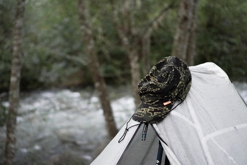 A camouflaged hat is placed atop a light gray tent in a wooded area with a river visible in the background. The scene conveys a sense of outdoor adventure and camping.