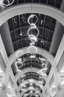 A series of illuminated circular light fixtures hang from the ceiling of a building with a modern architectural design. The ceiling features large archways and a glass paneling that provides a view of the lights from different angles.