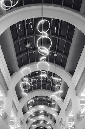 A series of illuminated circular light fixtures hang from the ceiling of a building with a modern architectural design. The ceiling features large archways and a glass paneling that provides a view of the lights from different angles.