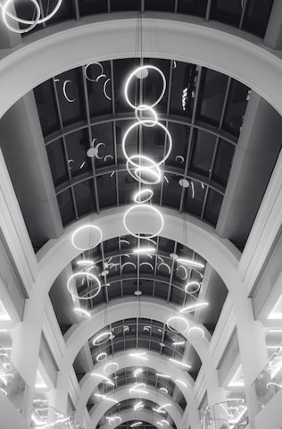 A series of illuminated circular light fixtures hang from the ceiling of a building with a modern architectural design. The ceiling features large archways and a glass paneling that provides a view of the lights from different angles.