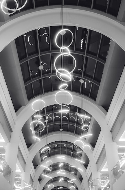 A series of illuminated circular light fixtures hang from the ceiling of a building with a modern architectural design. The ceiling features large archways and a glass paneling that provides a view of the lights from different angles.