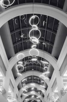 A series of illuminated circular light fixtures hang from the ceiling of a building with a modern architectural design. The ceiling features large archways and a glass paneling that provides a view of the lights from different angles.