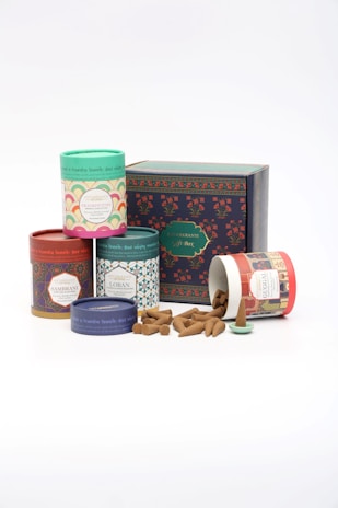 Assorted incense cones are displayed alongside colorful cylindrical packaging with intricate designs. A larger box labeled as a gift box is positioned behind the containers, adding a decorative and festive element.