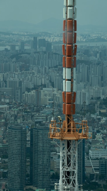 A large communication tower with a mix of red, white, and metal components stands prominently in the foreground. The backdrop features a sprawling urban landscape with densely packed high-rise buildings, indicating a metropolitan area. The sky is overcast, contributing to a muted color palette in the scene.