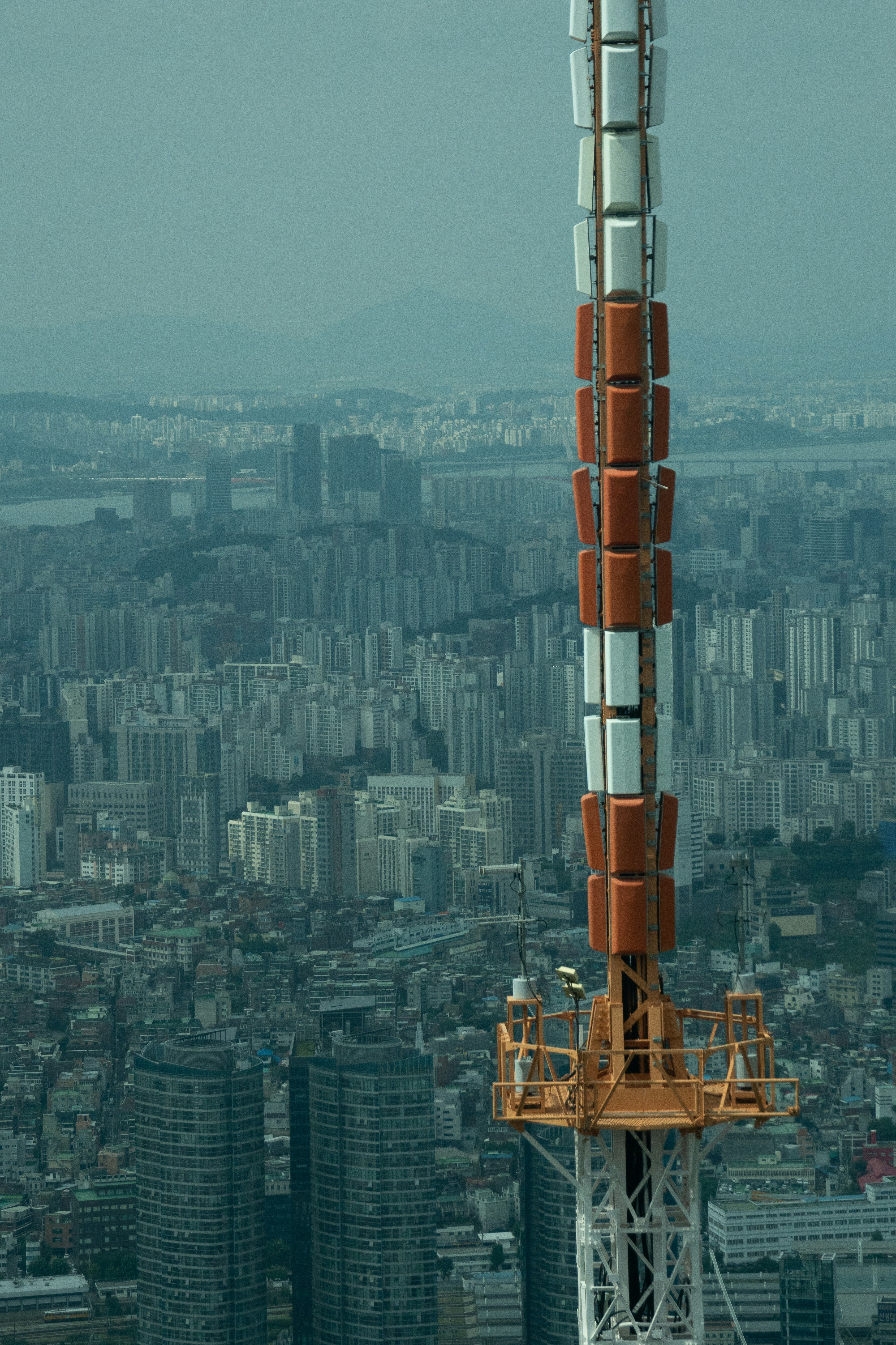 A large telecommunications or broadcast tower rises prominently against a sprawling urban skyline. The cityscape reveals numerous high-rise residential and commercial buildings, stretching toward distant mountains under a hazy blue sky. The tower's structure combines sections of orange, white, and grey metal, towering over the dense metropolitan area.