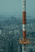 A large telecommunications or broadcast tower rises prominently against a sprawling urban skyline. The cityscape reveals numerous high-rise residential and commercial buildings, stretching toward distant mountains under a hazy blue sky. The tower's structure combines sections of orange, white, and grey metal, towering over the dense metropolitan area.