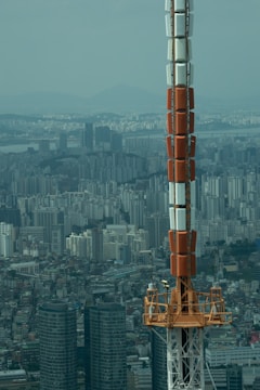 A large telecommunications or broadcast tower rises prominently against a sprawling urban skyline. The cityscape reveals numerous high-rise residential and commercial buildings, stretching toward distant mountains under a hazy blue sky. The tower's structure combines sections of orange, white, and grey metal, towering over the dense metropolitan area.