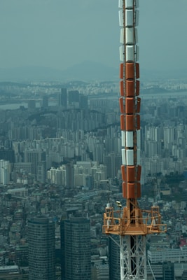 A large telecommunications or broadcast tower rises prominently against a sprawling urban skyline. The cityscape reveals numerous high-rise residential and commercial buildings, stretching toward distant mountains under a hazy blue sky. The tower's structure combines sections of orange, white, and grey metal, towering over the dense metropolitan area.