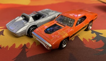 Two toy cars are positioned on a surface with a bright pattern. One is a sleek silver sports car with a convertible design, while the other is an orange muscle car featuring black stripes and chrome details. Both cars have a well-defined, glossy finish.