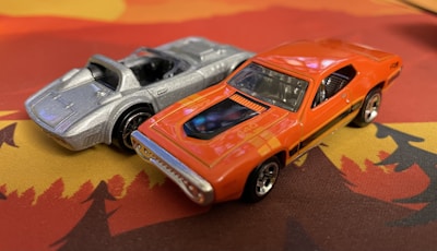 Two toy cars are positioned on a surface with a bright pattern. One is a sleek silver sports car with a convertible design, while the other is an orange muscle car featuring black stripes and chrome details. Both cars have a well-defined, glossy finish.