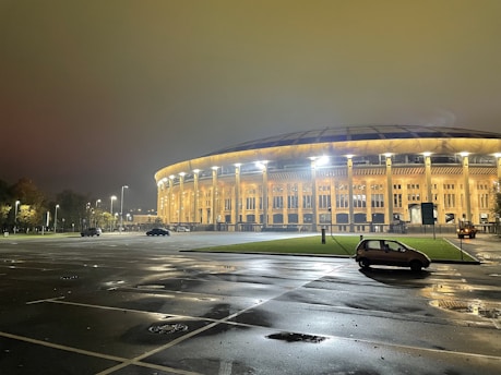A large, dome-shaped stadium illuminated with bright lights, creating a warm glow. The structure features tall pillars and numerous windows, with a mostly empty parking lot in the foreground. A few trees line the area, and two parked cars are visible near the stadium.