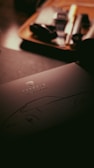 A dark, moody image featuring a document or brochure with the Porsche logo and the words 'Classic Register' on it. The background contains blurred items that appear to be on a wooden tray, suggesting a setting within an office or study.