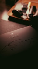 A dark, moody image featuring a document or brochure with the Porsche logo and the words 'Classic Register' on it. The background contains blurred items that appear to be on a wooden tray, suggesting a setting within an office or study.