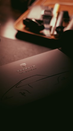 A dark, moody image featuring a document or brochure with the Porsche logo and the words 'Classic Register' on it. The background contains blurred items that appear to be on a wooden tray, suggesting a setting within an office or study.