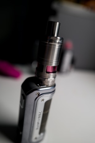 A close-up image of a vaping device with a metal tank and a sleek design. The surface is glossy and metallic, with visible screw details. A blurred background suggests an indoor setting with dim lighting.