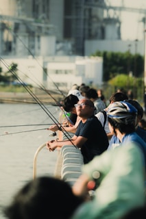 Several people are lined up along a railing by a body of water, engaged in fishing. Each person holds a fishing rod, with the lines cast into the water. The background shows an industrial or urban setting with buildings and greenery. The scene captures both leisure and concentration.