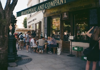 A group of people are gathered outside a quaint bookstore named Shakespeare and Company. They are seated on stools and chairs, engaged in conversation, with some people browsing books outside. The bookstore has a vintage and charming facade, with wooden elements and chalkboard signs. There is a canopy providing shade in part of the scene, and a tree partially frames the left side of the image.