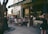 A group of people are gathered outside a quaint bookstore named Shakespeare and Company. They are seated on stools and chairs, engaged in conversation, with some people browsing books outside. The bookstore has a vintage and charming facade, with wooden elements and chalkboard signs. There is a canopy providing shade in part of the scene, and a tree partially frames the left side of the image.