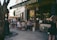 A group of people are gathered outside a quaint bookstore named Shakespeare and Company. They are seated on stools and chairs, engaged in conversation, with some people browsing books outside. The bookstore has a vintage and charming facade, with wooden elements and chalkboard signs. There is a canopy providing shade in part of the scene, and a tree partially frames the left side of the image.