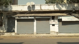A row of closed shopfronts with metal shutters down, situated in an older, possibly slightly run-down building. Above the shutters, there are signs in a local script and a couple of satellite dishes attached to the facade. Debris and dried leaves are scattered on the ground, and the building shows signs of wear and tear.
