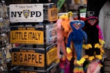 An assortment of souvenir license plates from New York with names of famous areas such as Little Italy and Big Apple is displayed on a rack. Nearby, colorful plush toys with exaggerated features, including bright colors and wide smiles, hang for sale. The setting appears to be a gift shop or a similar retail environment.