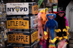 An assortment of souvenir license plates from New York with names of famous areas such as Little Italy and Big Apple is displayed on a rack. Nearby, colorful plush toys with exaggerated features, including bright colors and wide smiles, hang for sale. The setting appears to be a gift shop or a similar retail environment.