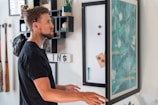A person with facial hair and a hearing aid is standing indoors, interacting with a large vertical board displaying abstract teal and white artwork. The surrounding decor includes shelves with assorted items like books and a baseball, as well as two baseball bats and a glove mounted on the wall.