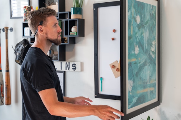 A person with facial hair and a hearing aid is standing indoors, interacting with a large vertical board displaying abstract teal and white artwork. The surrounding decor includes shelves with assorted items like books and a baseball, as well as two baseball bats and a glove mounted on the wall.