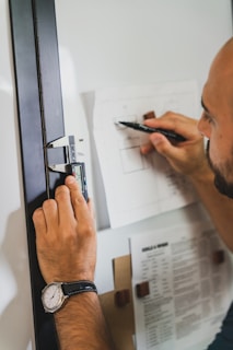 A person is using a digital caliper to take measurements on a piece of paper. The individual's left hand is holding the caliper, while the right hand is writing or drawing with a pen on the paper. The person is wearing a watch on their left wrist, and there are documents attached to a board nearby.