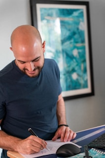 A person with a shaved head is standing at a desk, smiling and writing on a piece of paper with a pen. They are casually dressed in a dark t-shirt. On the desk, there is a computer mouse and a keyboard. A colorful abstract painting is hung on the wall in the background.
