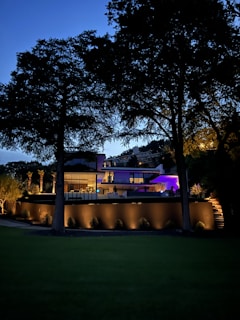 A modern, well-lit house is surrounded by tall trees during dusk. The structure features large glass windows and a blend of concrete and wooden elements. Soft lights illuminate the walls and landscape, creating a serene and inviting atmosphere. The sky is transitioning into night, adding a deep blue background.