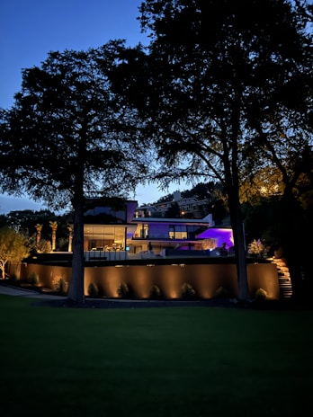 A modern, well-lit house is surrounded by tall trees during dusk. The structure features large glass windows and a blend of concrete and wooden elements. Soft lights illuminate the walls and landscape, creating a serene and inviting atmosphere. The sky is transitioning into night, adding a deep blue background.