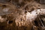 An expansive underground cave with a variety of stalactites hanging from the ceiling and stalagmites rising from the ground. The cave interior is illuminated, highlighting the rock formations. The walls and ceiling have intricate, natural patterns formed over time.