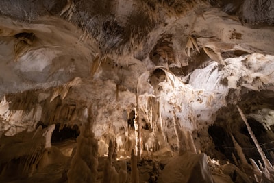 An expansive underground cave with a variety of stalactites hanging from the ceiling and stalagmites rising from the ground. The cave interior is illuminated, highlighting the rock formations. The walls and ceiling have intricate, natural patterns formed over time.