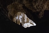 A dark cave with rugged, textured walls. A patch of light illuminates part of the cave, highlighting the rough surface and variations in color and shadow. Stalactites and various formations are partially visible, hanging from the cave ceiling.