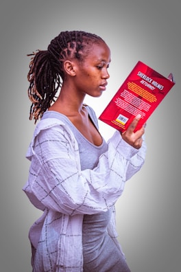 A person with braided hair is intently reading a book titled 'Sherlock Holmes Returns.' They are wearing a grey dress and a light-colored, patterned shirt. The focus is on the book and the reader, with a plain, gradient background adding a simple backdrop to the scene.