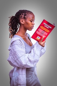 A person with braided hair is intently reading a book titled 'Sherlock Holmes Returns.' They are wearing a grey dress and a light-colored, patterned shirt. The focus is on the book and the reader, with a plain, gradient background adding a simple backdrop to the scene.
