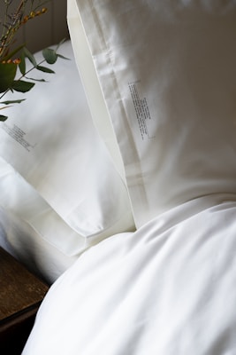 Two white pillows placed on a bed with a crisp white sheet. There is a small branch with green leaves and yellow berries partially visible on the left side.