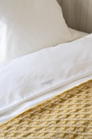 A neatly made bed featuring a white pillow and sheets, accented by a patterned yellow and white knitted throw blanket. The setting conveys a sense of cleanliness and comfort.