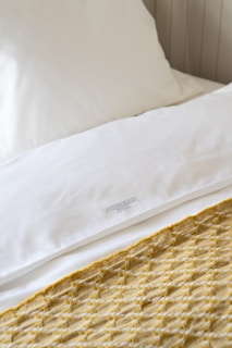 A neatly made bed featuring a white pillow and sheets, accented by a patterned yellow and white knitted throw blanket. The setting conveys a sense of cleanliness and comfort.