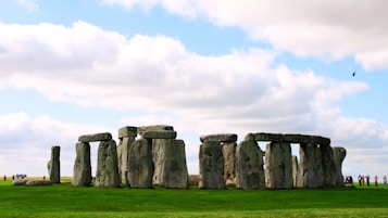 A prehistoric monument consisting of massive stone structures arranged in a circular layout, set against a backdrop of a partly cloudy sky. People are visible in the distance, standing and taking photographs.