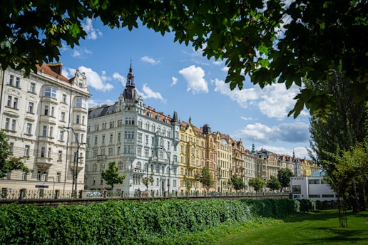 A row of ornate, historic buildings with diverse architectural styles lines a tree-covered street. The sky is bright with scattered clouds, and a lush green hedge runs alongside the buildings.